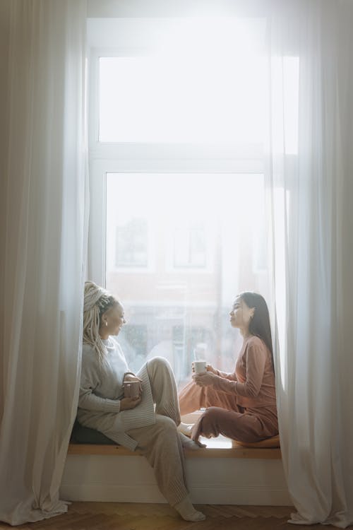 Free Women Sitting by the Window while Looking at Each Other Stock Photo