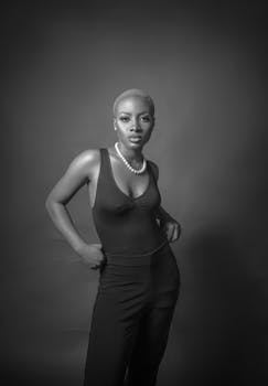 A confident woman in a black bodysuit and pearl necklace posing in a studio.