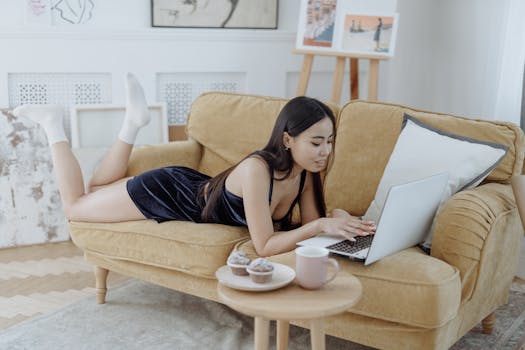 Asian woman in a cozy home setting, browsing on a laptop while enjoying cupcakes and a mug of coffee.