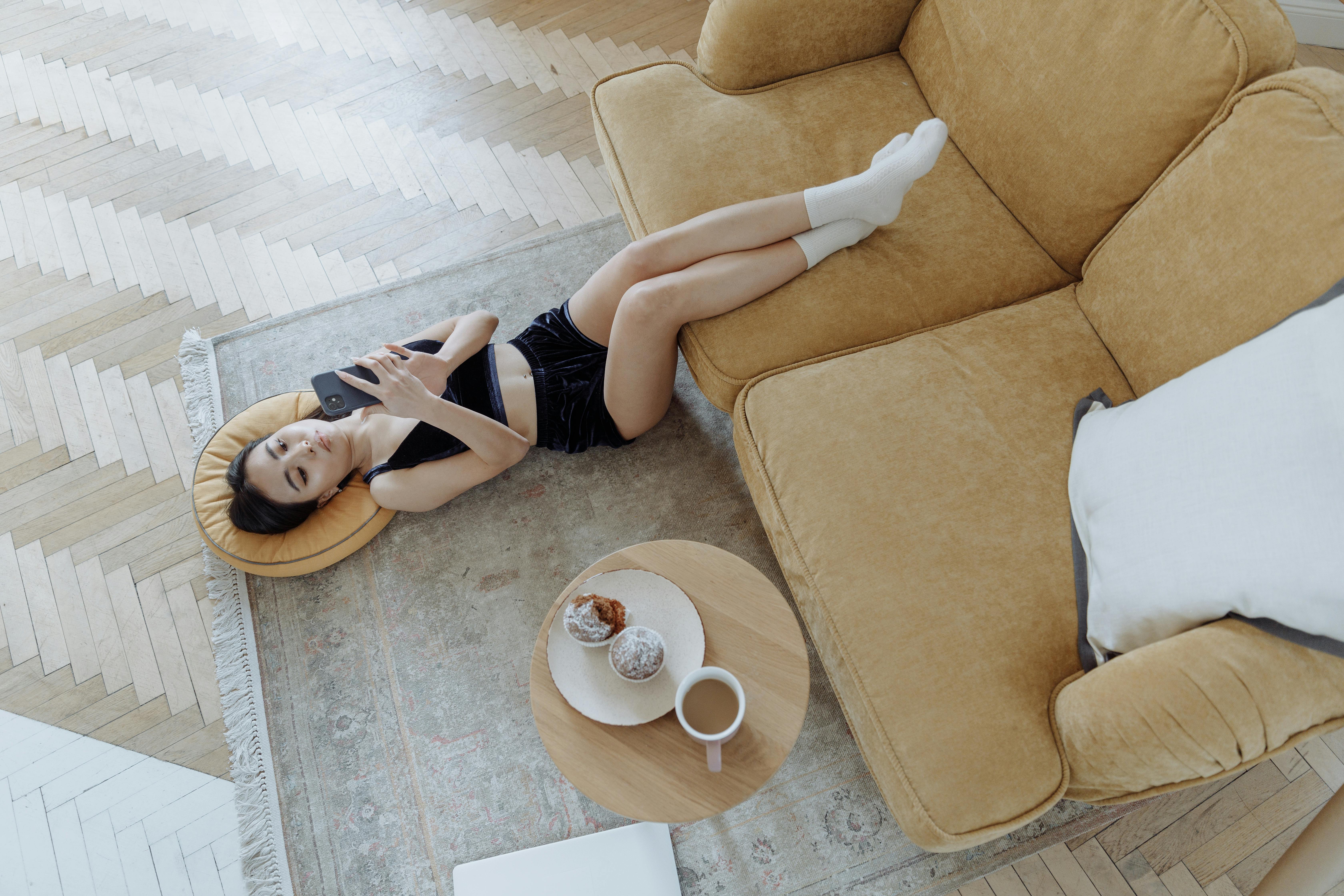 Woman lying on carpet using cellphone with snack and coffee nearby, enjoying a relaxing moment indoors.