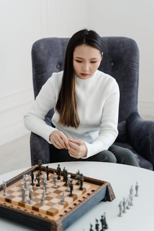 Woman in White Sweater Sitting on Single Sofa Playing Chess Game
