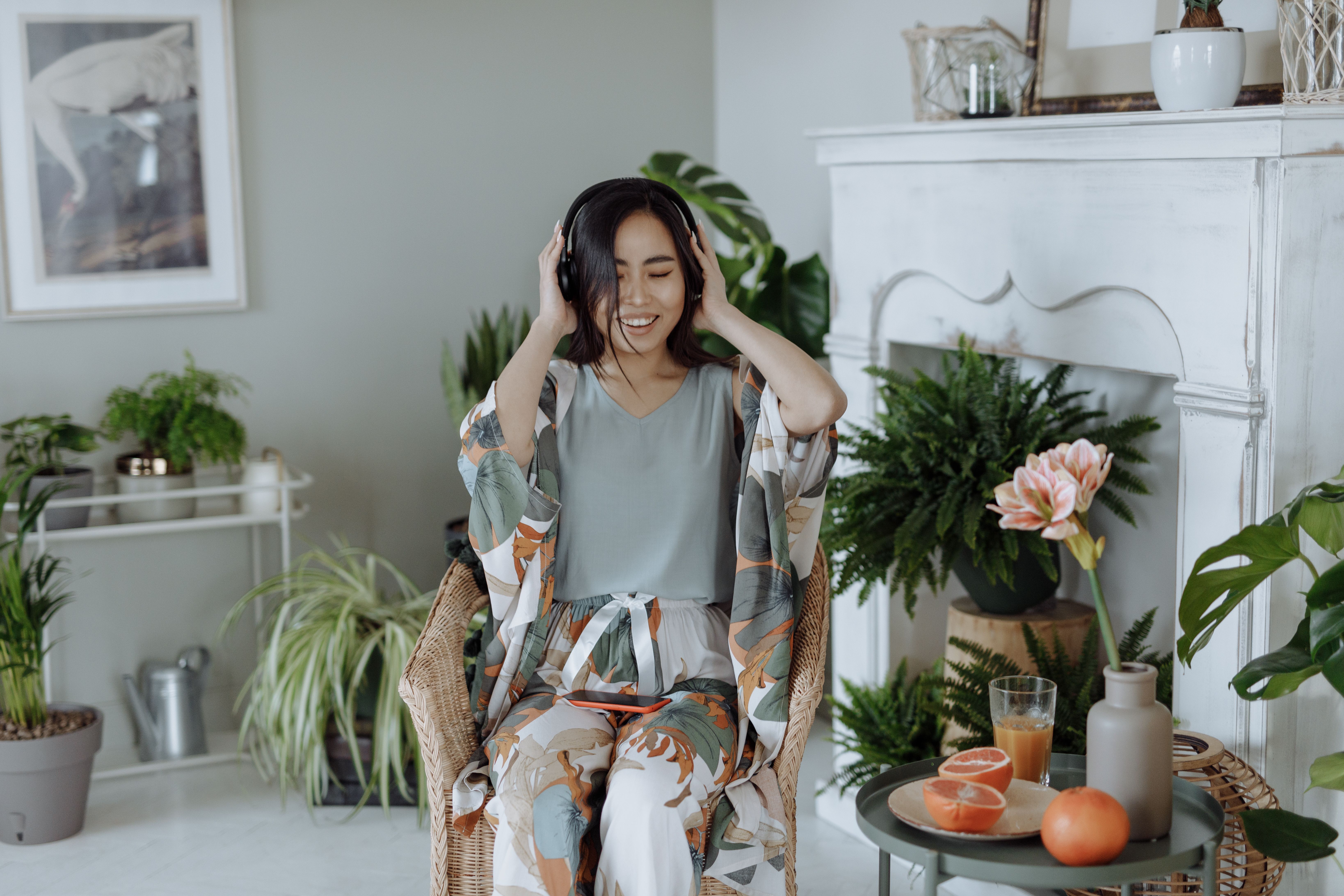 Relaxed Asian woman wearing pajamas and headphones, surrounded by lush indoor plants in a stylish home setting.