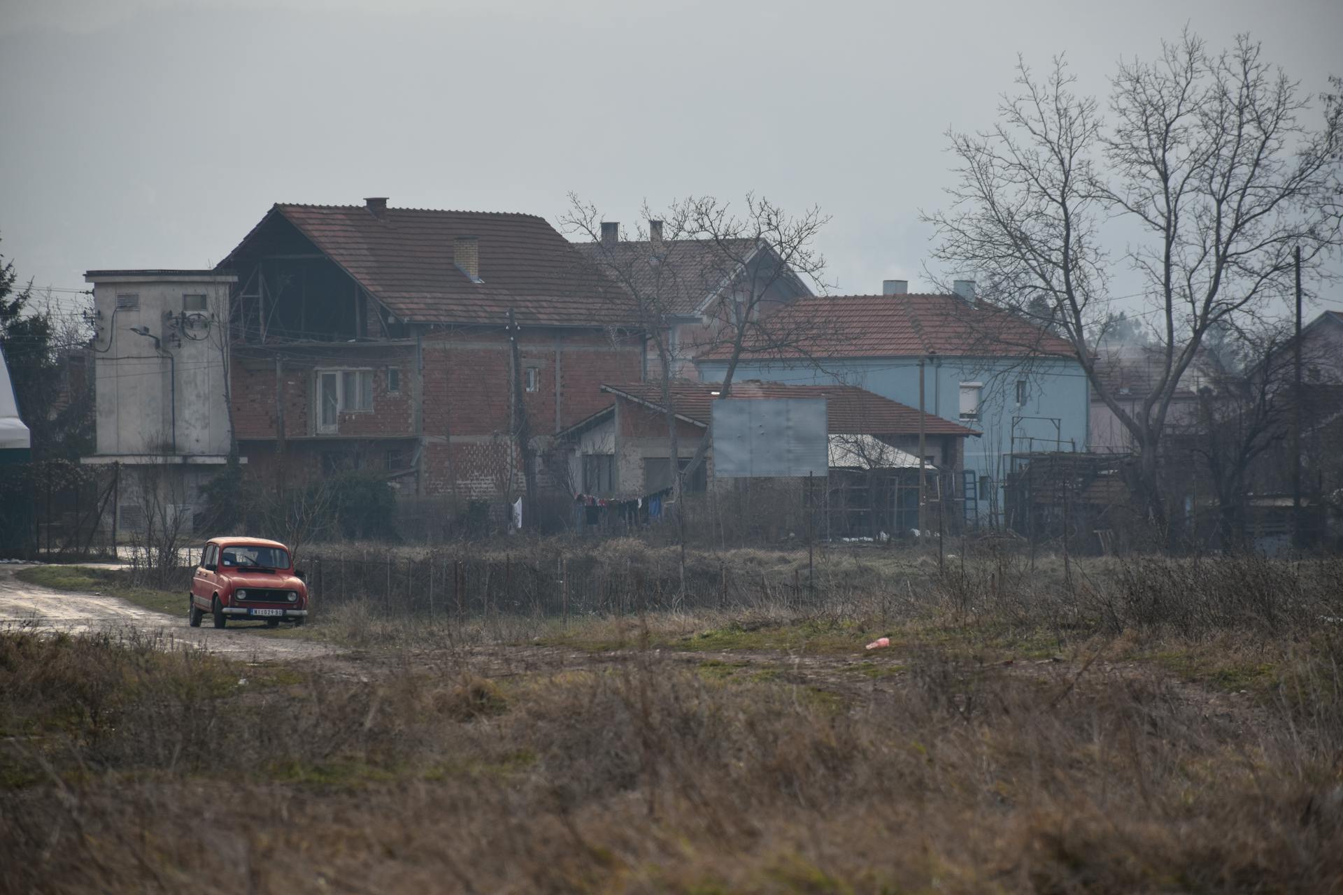 A rural scene featuring brick houses in disrepair and a vintage red car on a cloudy day.