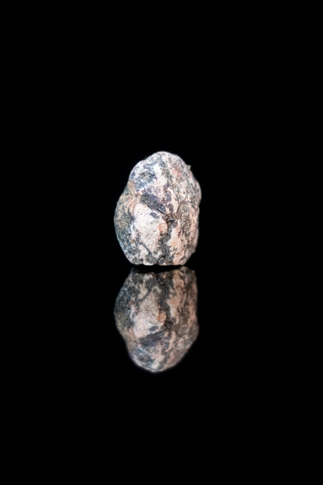 A single rough gray and cream speckled stone standing upright on a black reflective surface, lit from above with its mirror image below