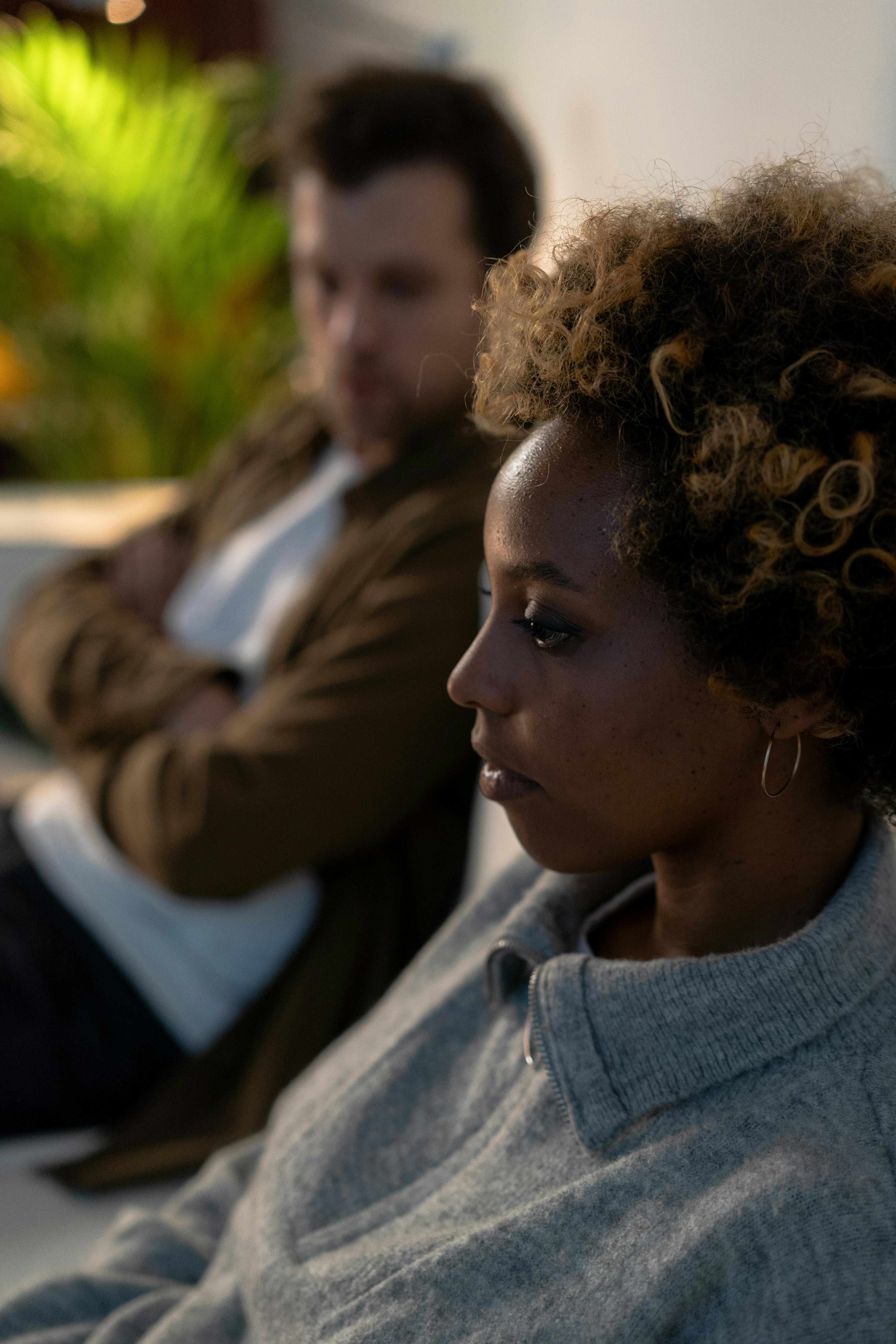 A woman with afro hair sits pensively on a sofa, captured in a cozy indoor setting.