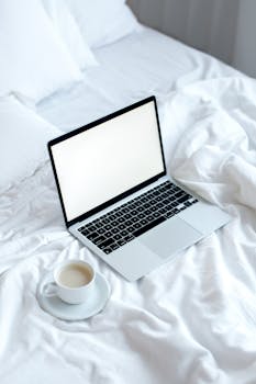 A laptop with a blank screen and a coffee cup on a neatly made bed, providing a serene and cozy workspace vibe.