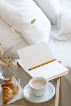 A serene morning setup with coffee, croissant, notepad, and pencil on a glass table by a bed.