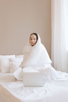 A woman wrapped in a white blanket sits on a bed using a laptop, indoors.