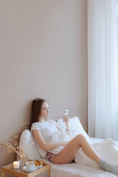 Young woman enjoys breakfast in bed, sipping coffee and using her phone in a peaceful morning setting.