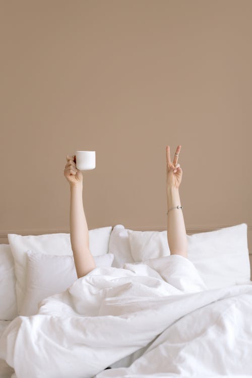 A Person Lying in Bed Holding Up a Cup of Drink Doing a Peace Sign