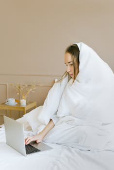 Woman wrapped in a white blanket using a laptop in a cozy bedroom setting, exuding comfort and relaxation.