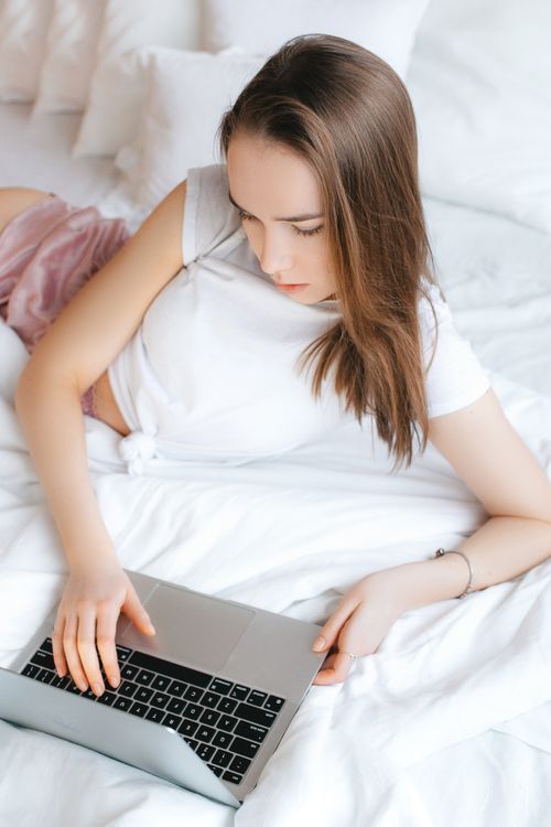 Free A young woman relaxing with a laptop on a white bed, embodying modern digital lifestyle and comfort. Stock Photo