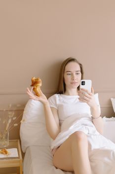 Woman enjoying a croissant while using her smartphone in bed, showcasing a cozy and relaxed morning routine.