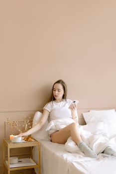 Young woman in white top enjoying a cozy moment on bed with smartphone in hand.