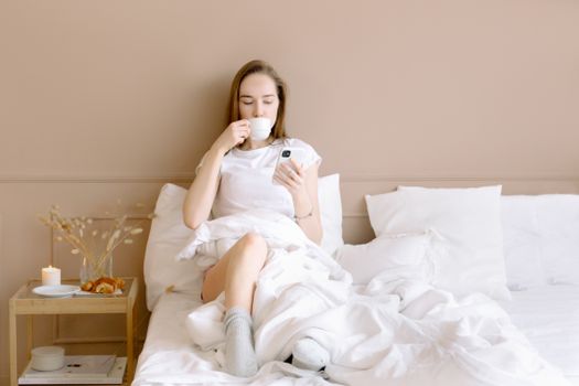 Relaxed woman in bed drinking coffee and using smartphone in a cozy setting.