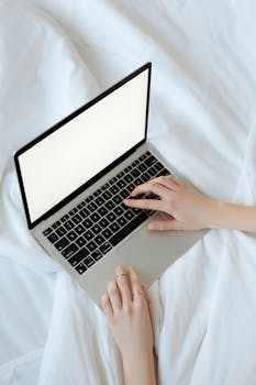 Close-up of hands typing on a laptop in a well-lit bedroom setting, featuring a clean minimalistic style.