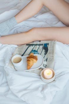 Relaxing morning scene with croissant and coffee in bed, accompanied by a candle and newspaper.