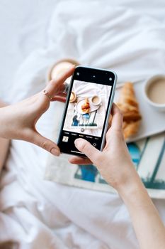 Hands holding smartphone to photograph coffee and croissant breakfast setup.