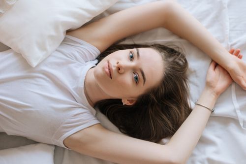 Free A woman lying down on her bed with white linens, in a relaxed pose wearing a white shirt. Stock Photo