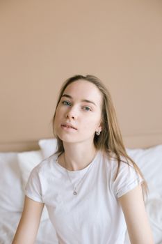 A serene portrait of a young Caucasian woman sitting on a bed in a softly lit room.