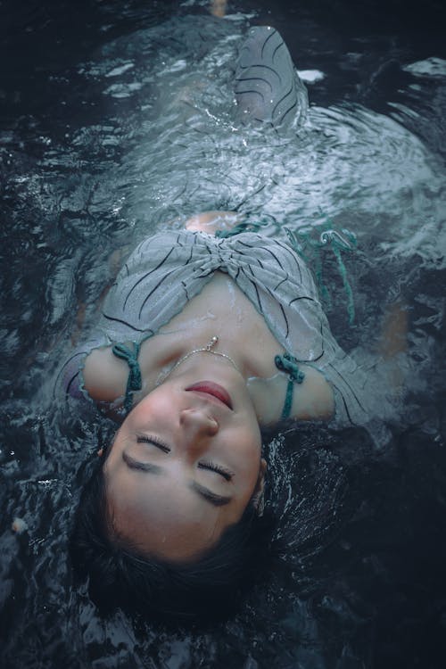 From above of serene female with closed eyes lying in transparent water of creek in nature