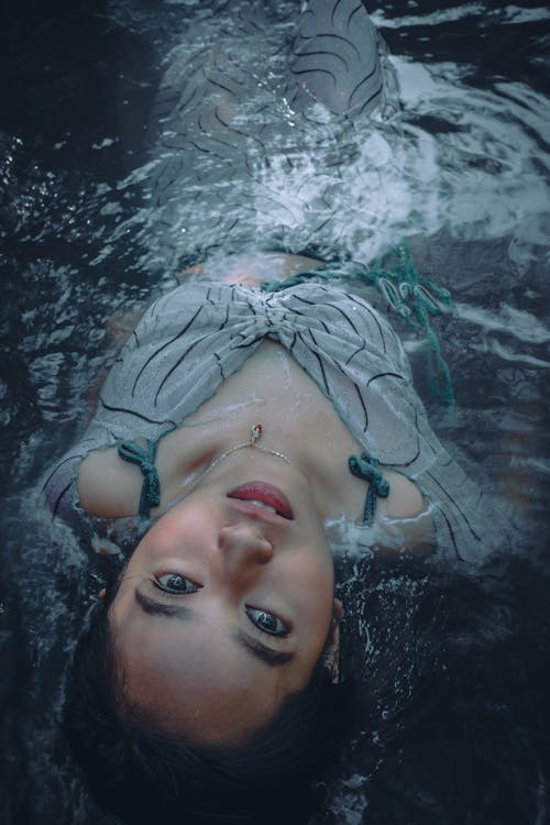 From above of peaceful Asian female looking at camera while lying in transparent water of river while resting in nature