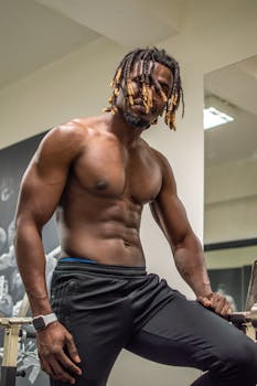 Muscular black man with dreadlocks poses shirtless in gym setting.