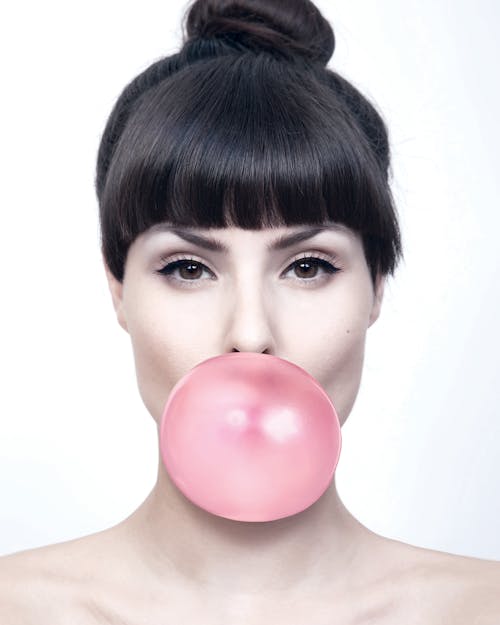 Free Young female model with makeup and bare shoulders blowing bubble gum against white background and looking at camera Stock Photo