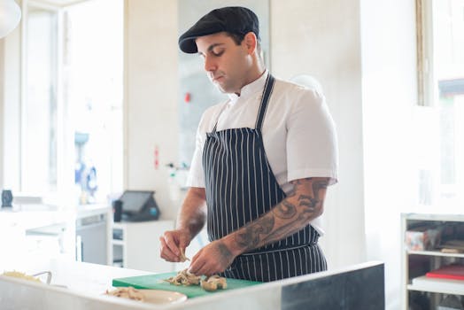 Tattooed chef cuts mushrooms on a chopping board in a bright, modern kitchen.