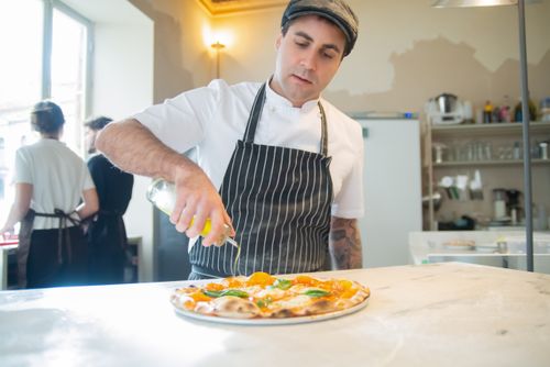 Free Chef drizzles olive oil on a freshly made pizza in a bright Italian kitchen setting. Stock Photo