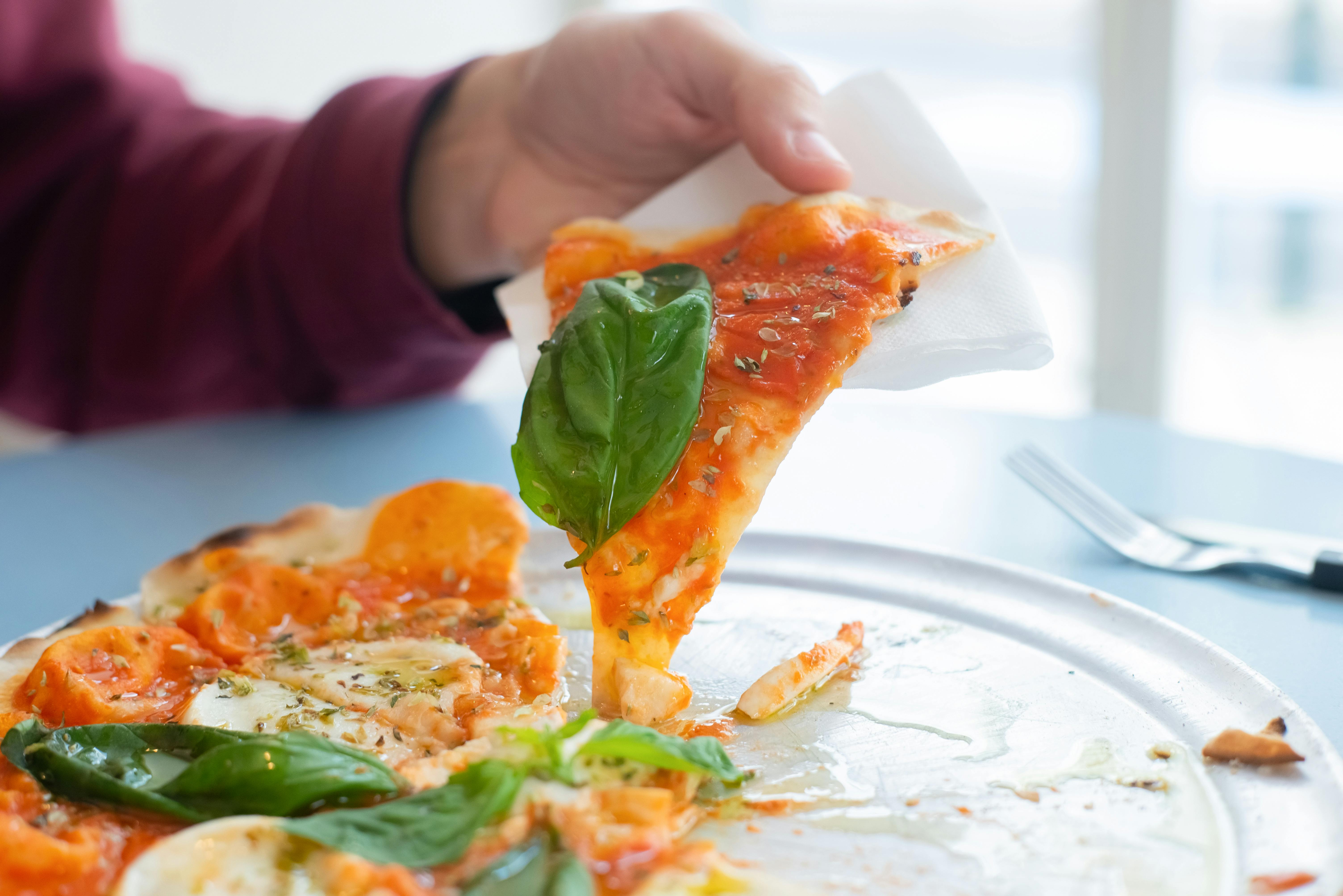 Close-up of a hand holding a slice of delicious Margherita pizza with basil topping on a sunny day. - Photo by Kampus Production on Pexels
