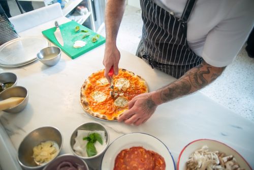 Free Chef slices delicious pizza with cheese in a modern kitchen. Fresh ingredients surround the dish. Stock Photo
