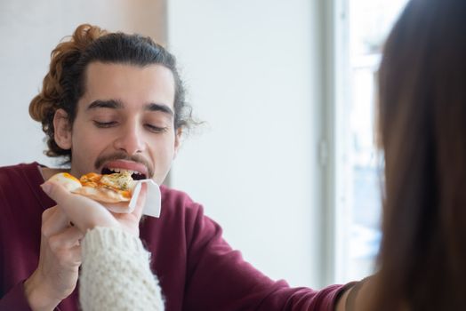 A man eagerly taking a bite of pizza offered by a friend in a cozy indoor setting.