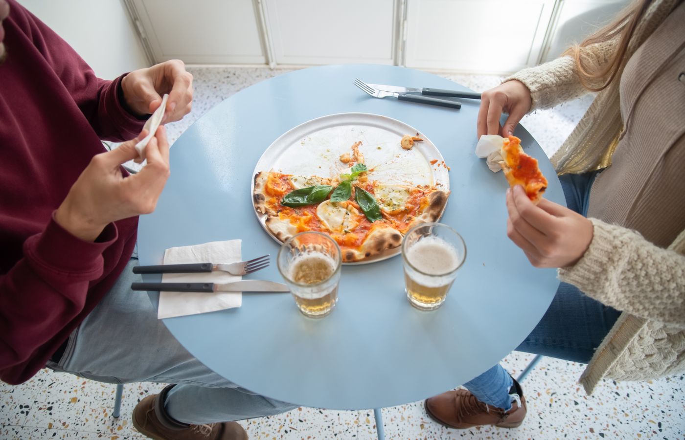Overhead view of friends sharing pizza and beer at a table indoors in a casual setting.