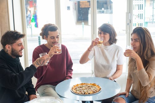 A Group of People Sitting at a Table with a Tray of Pizza Holding Glasses of Drinks
