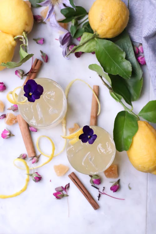 Top view of glasses with tasty alcohol beverages with ice decorated with flowers placed on table with cinnamon and lemons