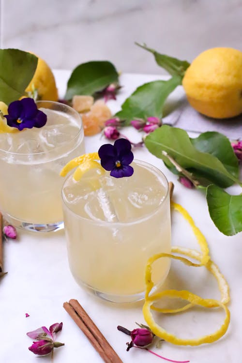 From above of transparent glasses with alcohol drinks and ice decorated with flowers and zest placed on table with ripe lemons and cinnamon