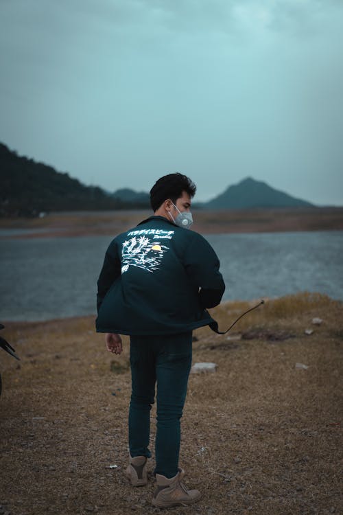 Man in Black Jacket Walking on Brown Grass in Front of a River