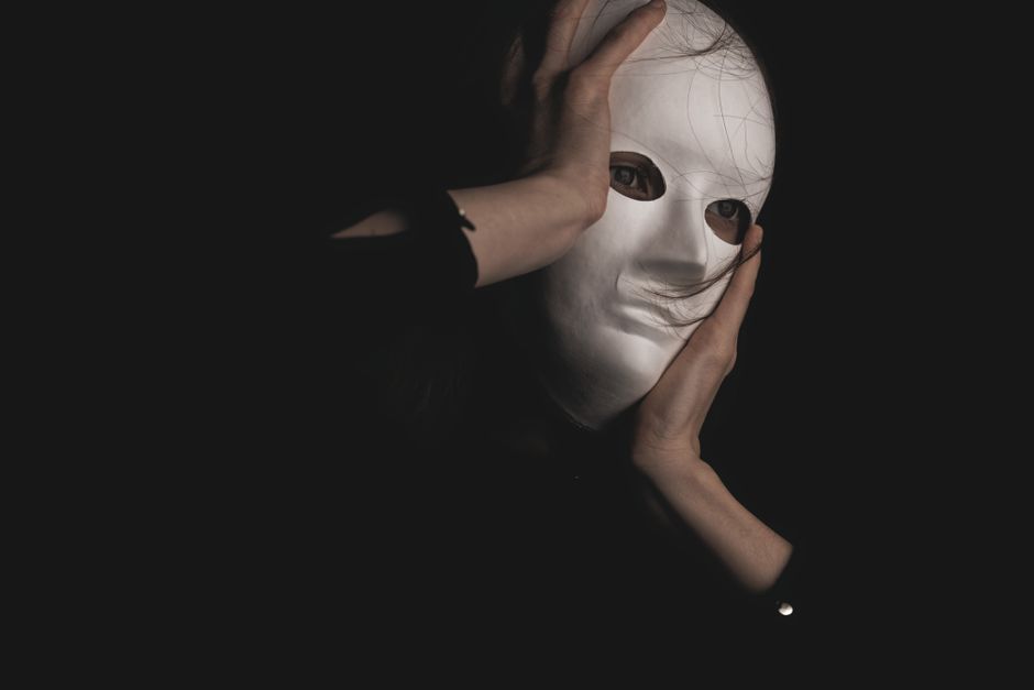 Close-up of a person holding a white mask against a dark background, symbolizing mystery.