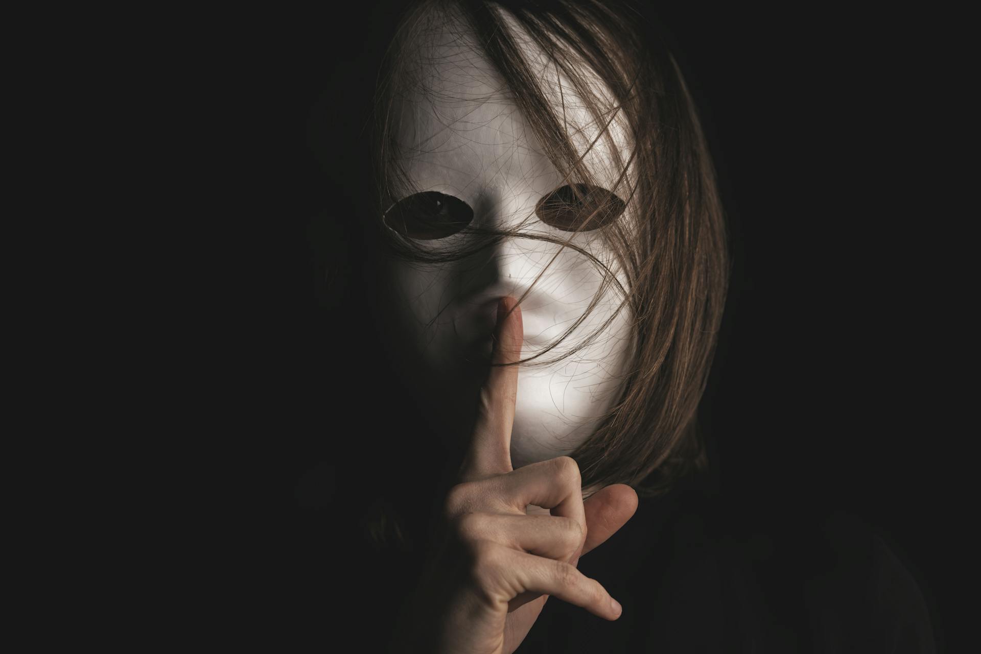 A person with a white mask makes a silence gesture, conveying mystery and secrecy.