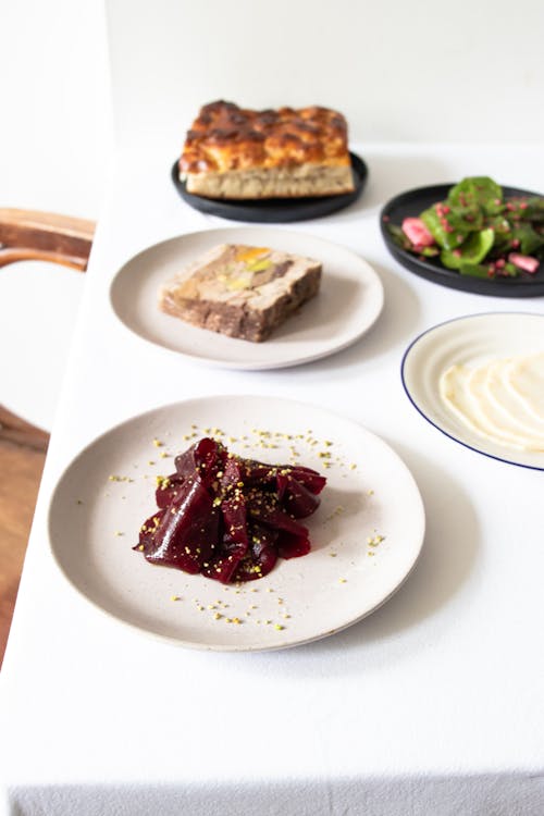 From above plates of beetroot dish and meatloaf served on white table with fresh salad and bread in light restaurant