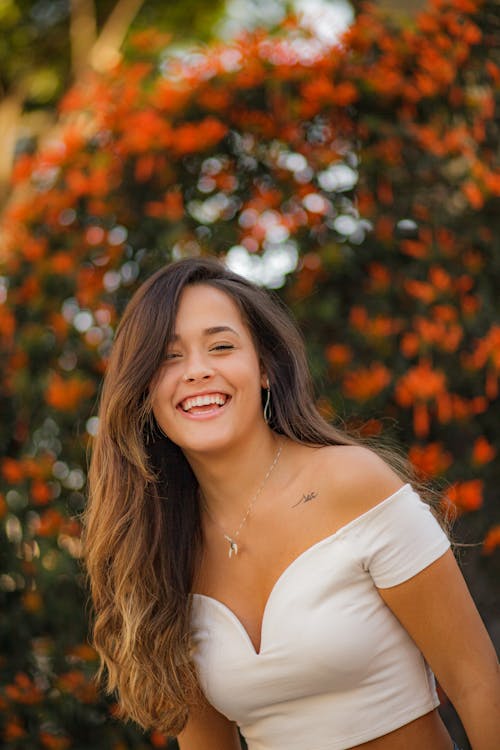 Laughing female in stylish outfit smiling and looking at camera while standing near blooming trees in garden on blurred background