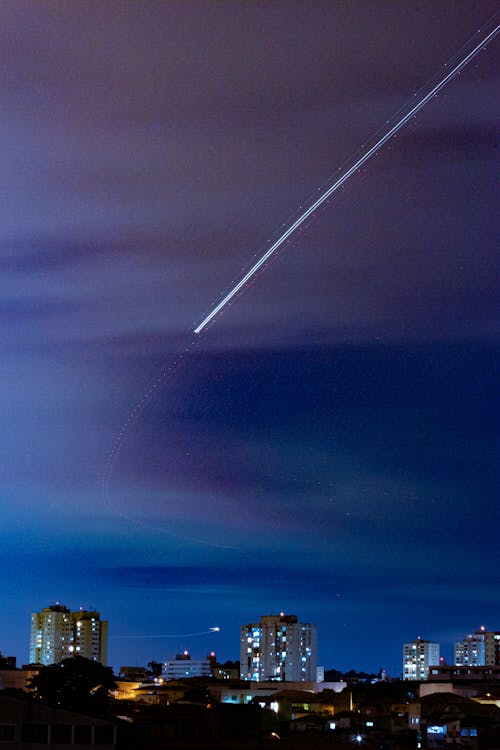 Bright shiny trail of plane flight above city with luminous tall buildings at late night under dark blue cloudy sky