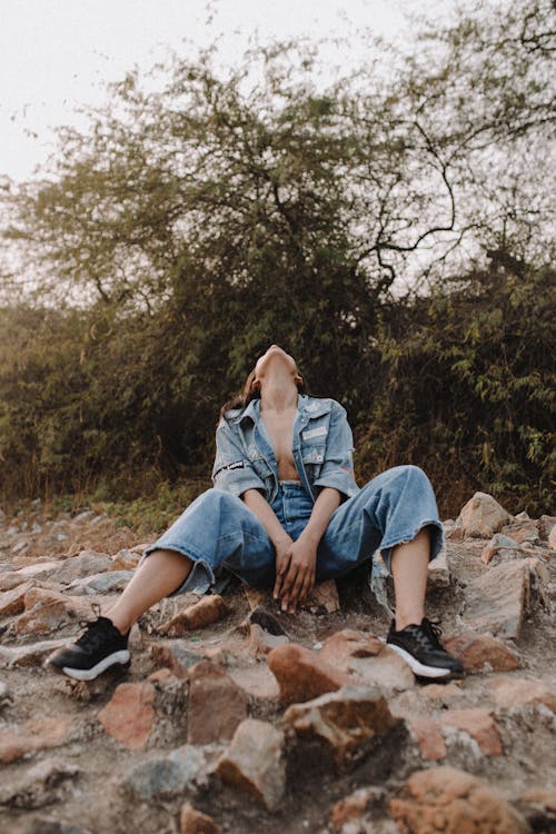 Full length of faceless female in casual clothes sitting on stony terrain near green trees and plants in daylight in nature