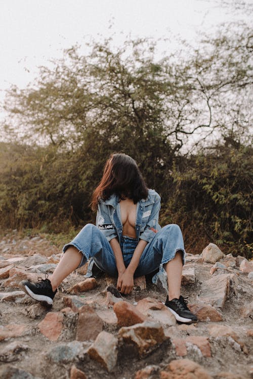 Full length of faceless female in casual clothes sitting on stony terrain near green trees and plants with shrubs in daylight in nature