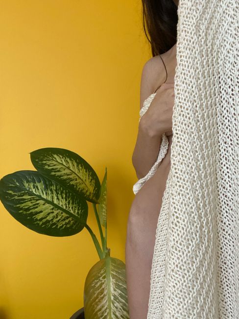 Unrecognizable nude slim female with dark hair hiding behind curtain while standing near potted houseplant with green leaves on yellow background