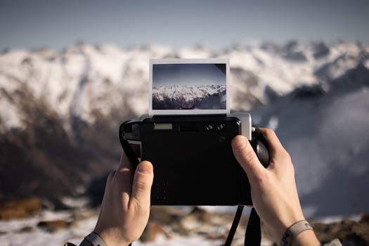 Person holding an instant camera capturing a scenic snow-capped mountain view.