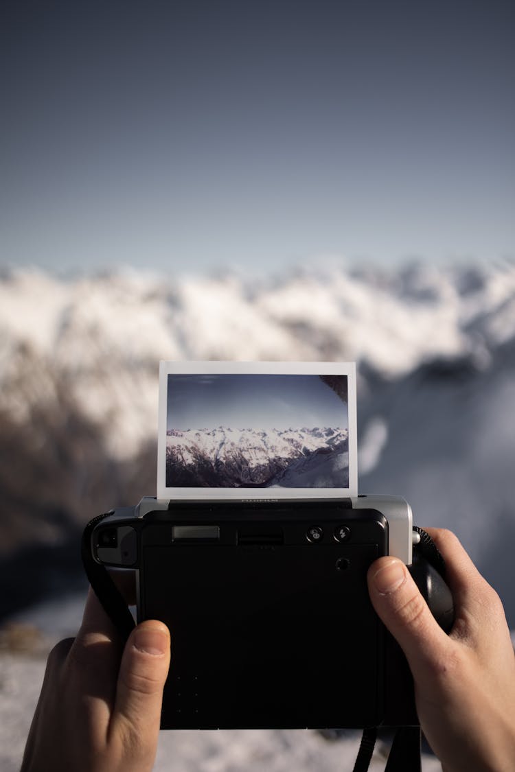 A Person Taking Picture Of A Snow Covered Mountain With A Polaroid Camera