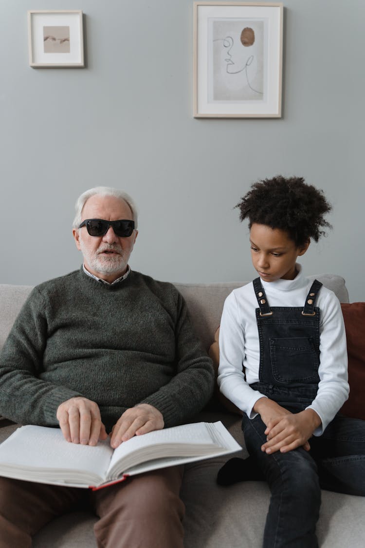 A Man Wearing Sunglasses Sitting On Sofa Reading A Braille Book Beside A Child