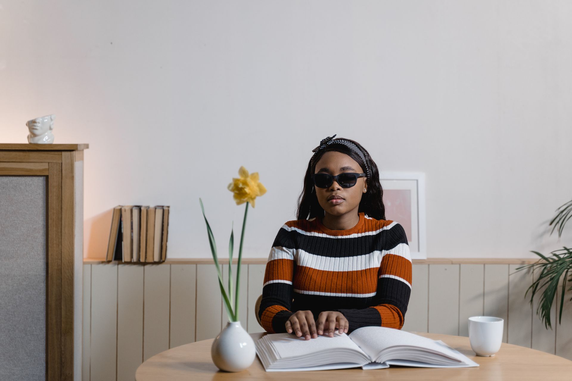 A blind woman in sunglasses reads a Braille book at a table with a flower vase indoors.